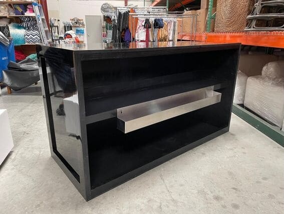 Black High Gloss Bar Table shop counter with shelving and a metal bar across the back is placed on a concrete floor in an industrial-looking retail space.
