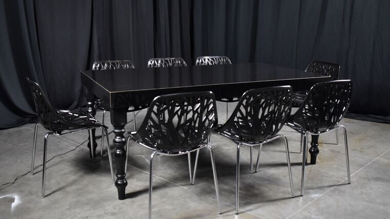 A modern, Black High Gloss Black Legs Dining with intricate-designed chairs is set against dark curtains on a concrete floor, creating a sleek, contemporary atmosphere.