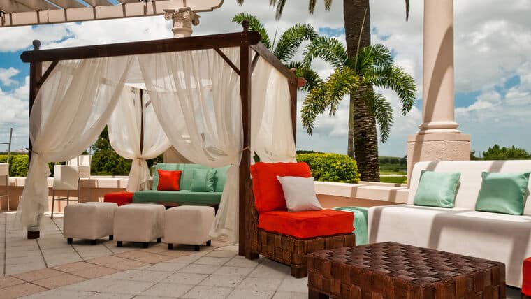 Outdoor patio with a cozy cabana featuring white drapes, vibrant seating with red and green cushions, palm trees, and a sunny sky. Relaxing atmosphere.