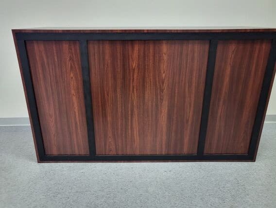 Mahogany Bar Wooden desk with a dark brown finish, featuring vertical panels framed in black. It sits on a light gray tiled floor against a plain wall.
