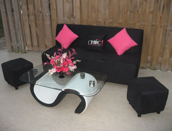 Outdoor seating area with a black sofa, two pink pillows, and a "Chic!" pillow. A glass coffee table holds a vibrant pink flower arrangement
