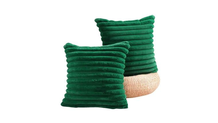 Two Green Ribbed Emerald Velvet pillows stand on a light-colored woven pouf against a white background, conveying a sense of comfort and modern style.