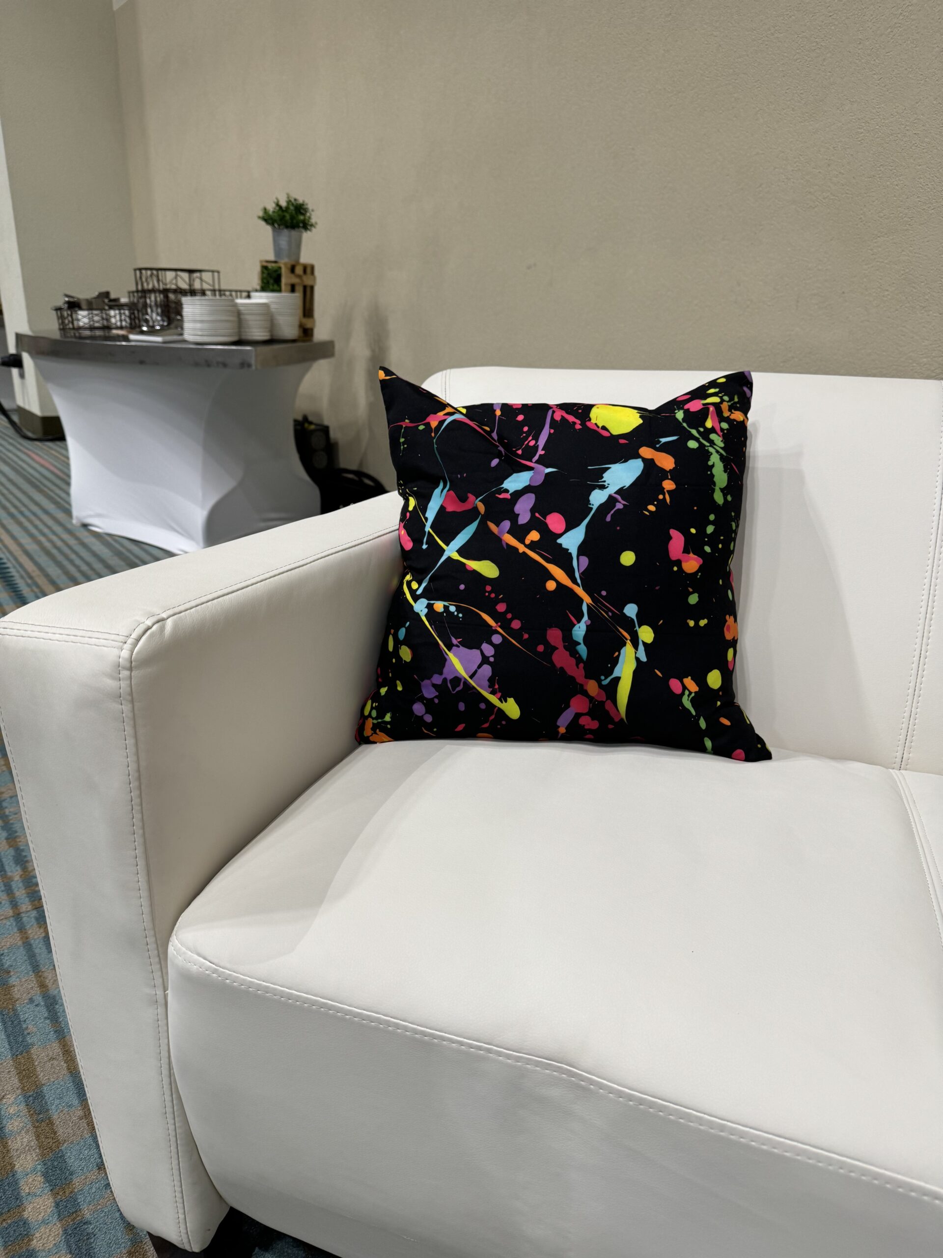 A white couch holds a black pillow with a vibrant paint splatter design.