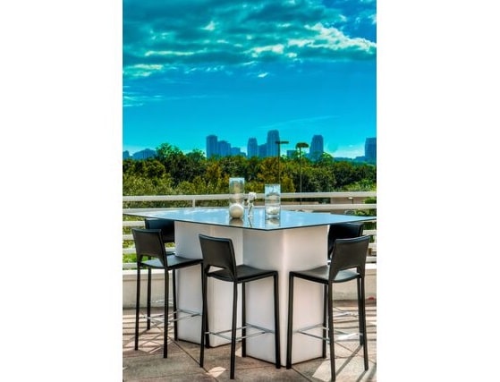 Modern outdoor patio with a white table and four black chairs on a sunny day. A city skyline and lush trees are visible under a bright blue sky.