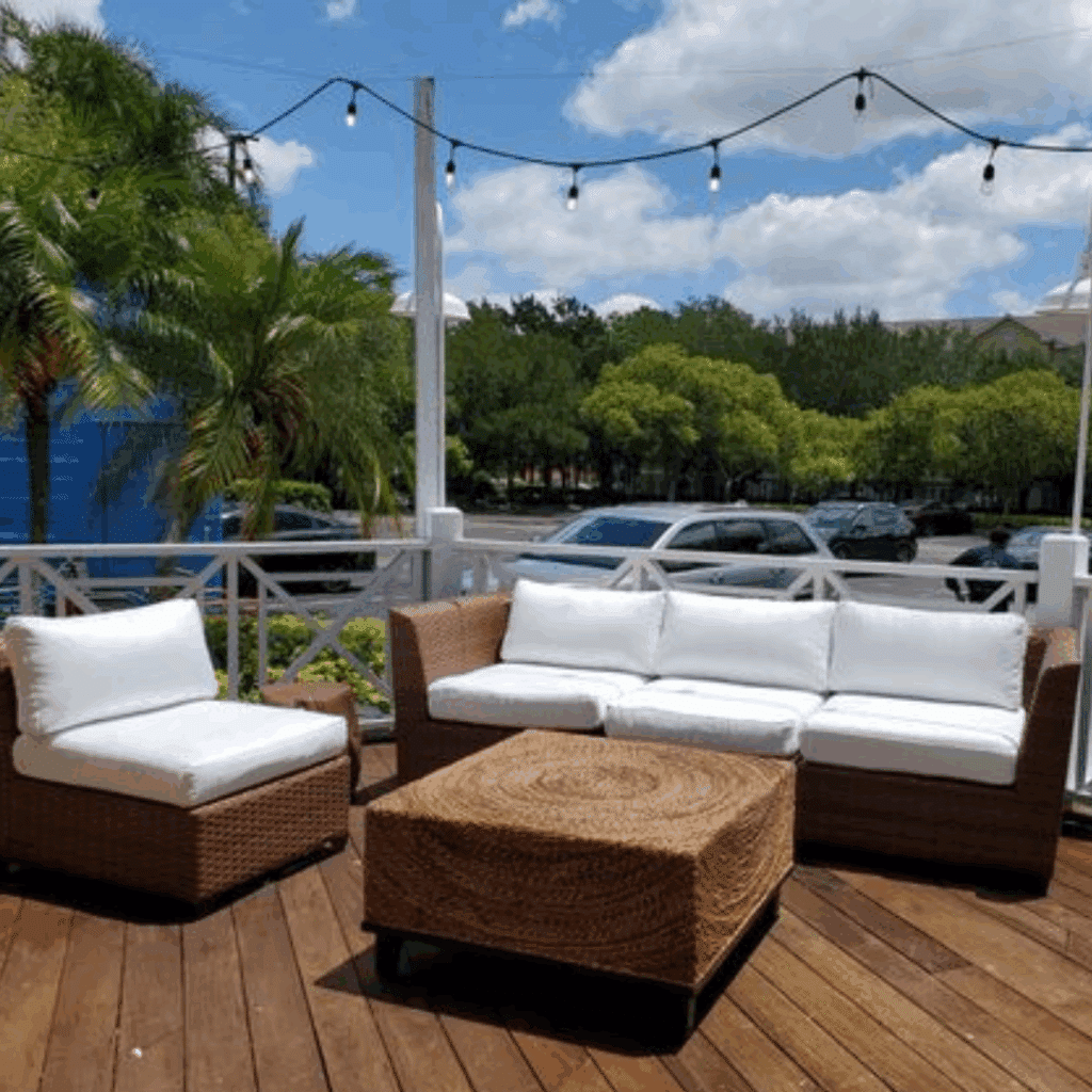 Captiva Corner Chair patio furniture with white cushions sits on a wooden deck. Palm trees and string lights create a relaxed, tropical atmosphere under a blue sky.