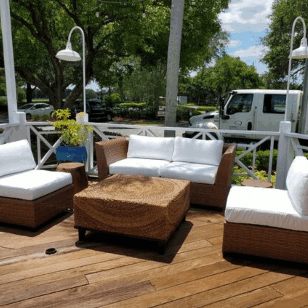 Captiva Loveseat Outdoor patio with wicker furniture and white cushions, surrounded by trees. A wicker coffee table sits in the center, under a clear blue sky.