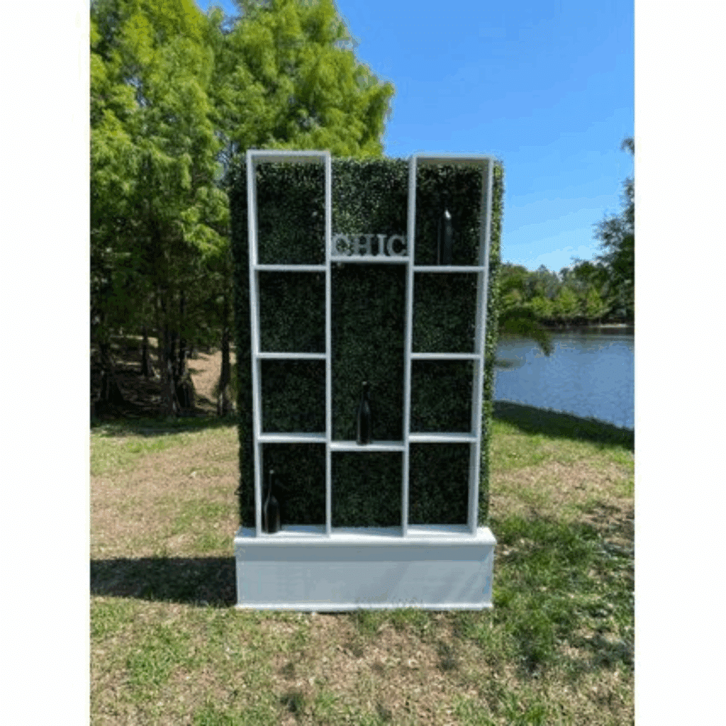 Hedge Wall Display Box shelving unit with green leafy backing labeled "CHIC," holding black bottles. Set outdoors by a peaceful lake and trees under blue sky.
