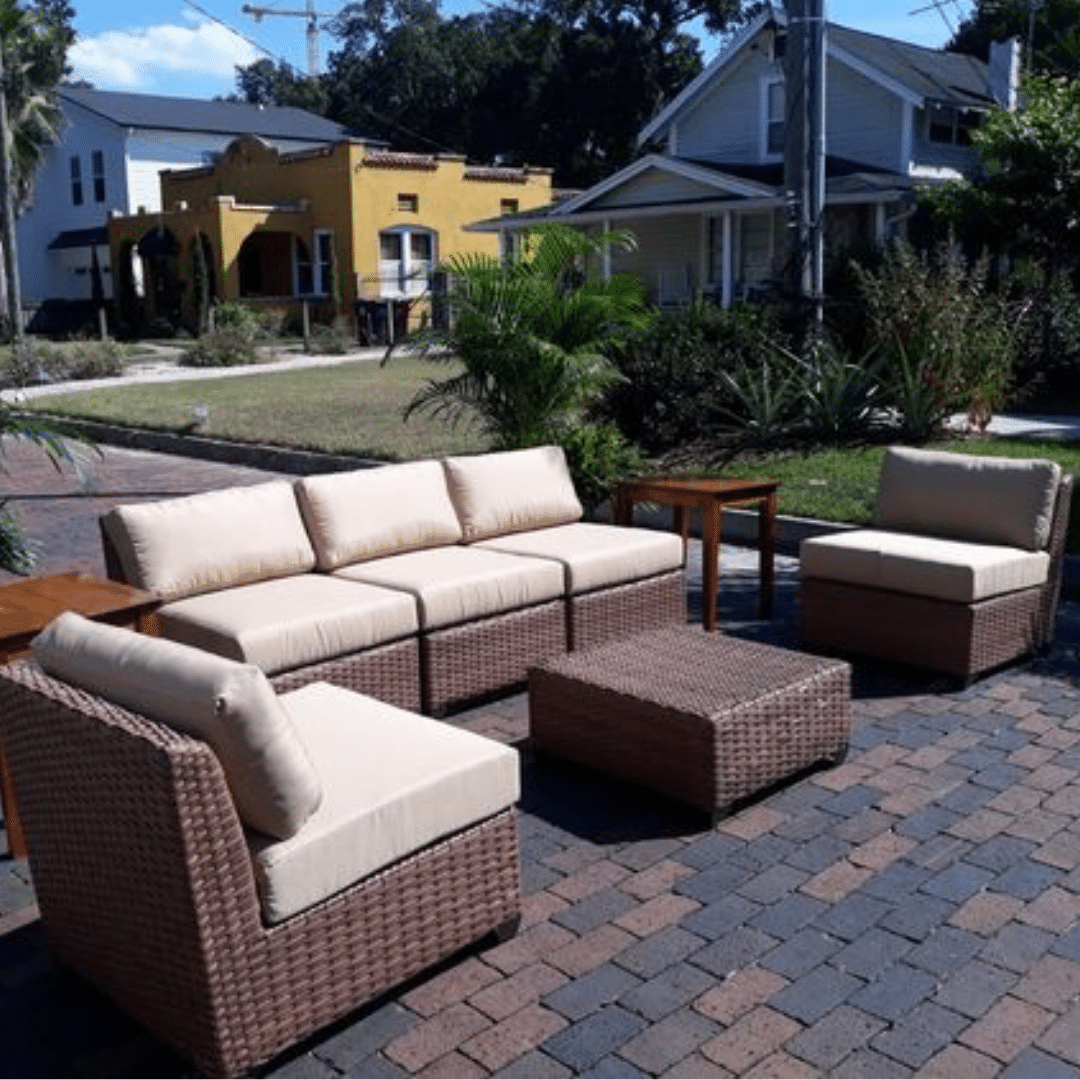 Outdoor Outdoor Event Furniture with beige cushions. The setup includes a sofa, chairs, and tables on a brick-paved patio. Plants and houses are visible in the background, creating a cozy, inviting atmosphere.