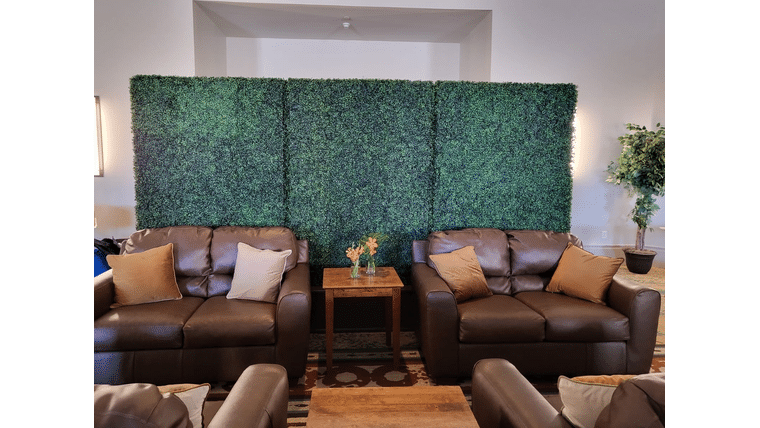 Standard Boxwood Walls with beige pillows face each other, separated by a small wooden table with flowers. A large green hedge spans the background, conveying a cozy yet fresh atmosphere.